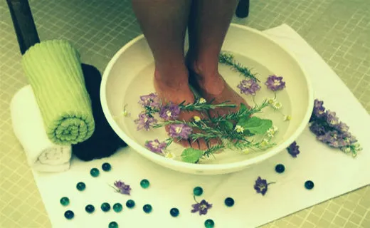 Pés mergulhados em uma bacia com água, ervas e flores lilases, ao lado de toalhas enroladas e bolinhas de gude