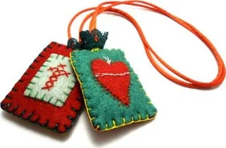 Dois patuás de feltro, um vermelho com bordado de cruzes e outro verde com um coração vermelho, presos a um cordão laranja.