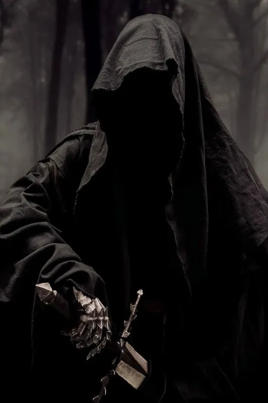 Figura encapuzada envolta em manto negro, com o rosto oculto na sombra, segurando um cajado de metal numa floresta enevoada.