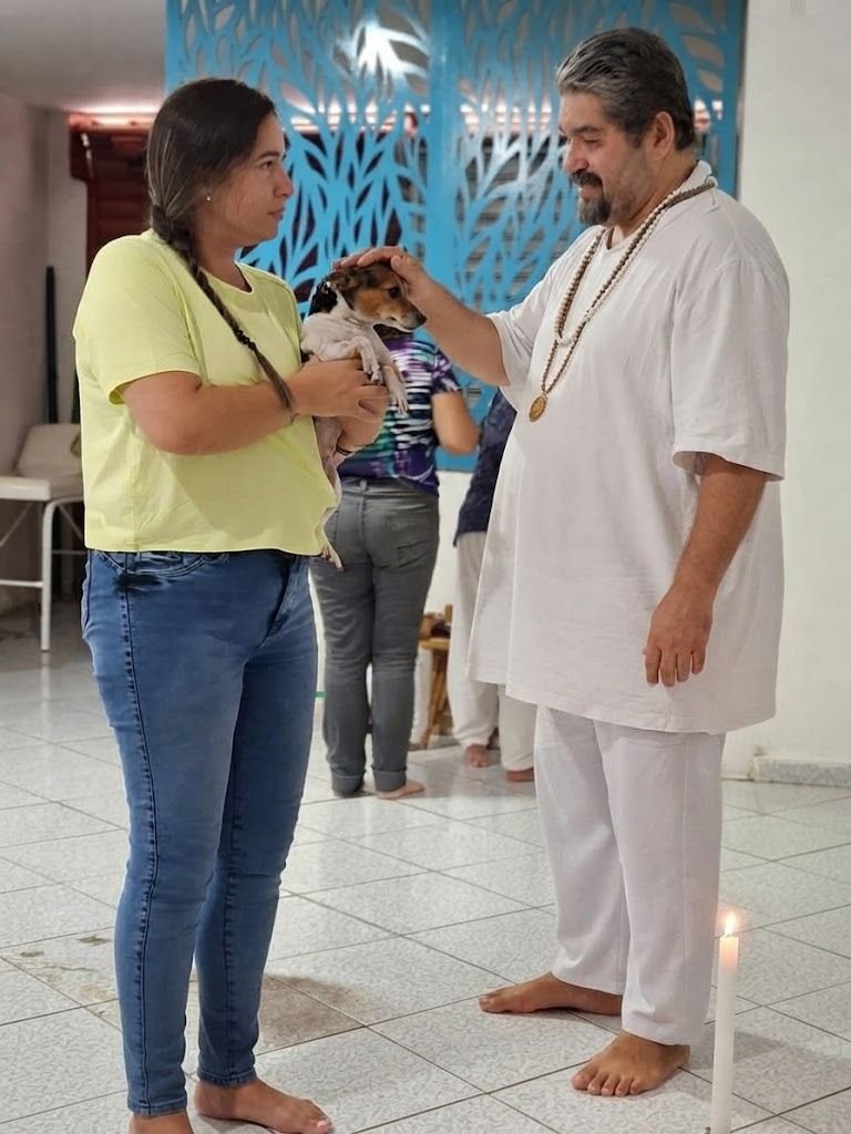 O dirigente do Templo, de branco e com guias, impõe a mão sobre a cabeça de um cão de pequeno porte no colo de sua tutora, ao lado de uma vela acesa.