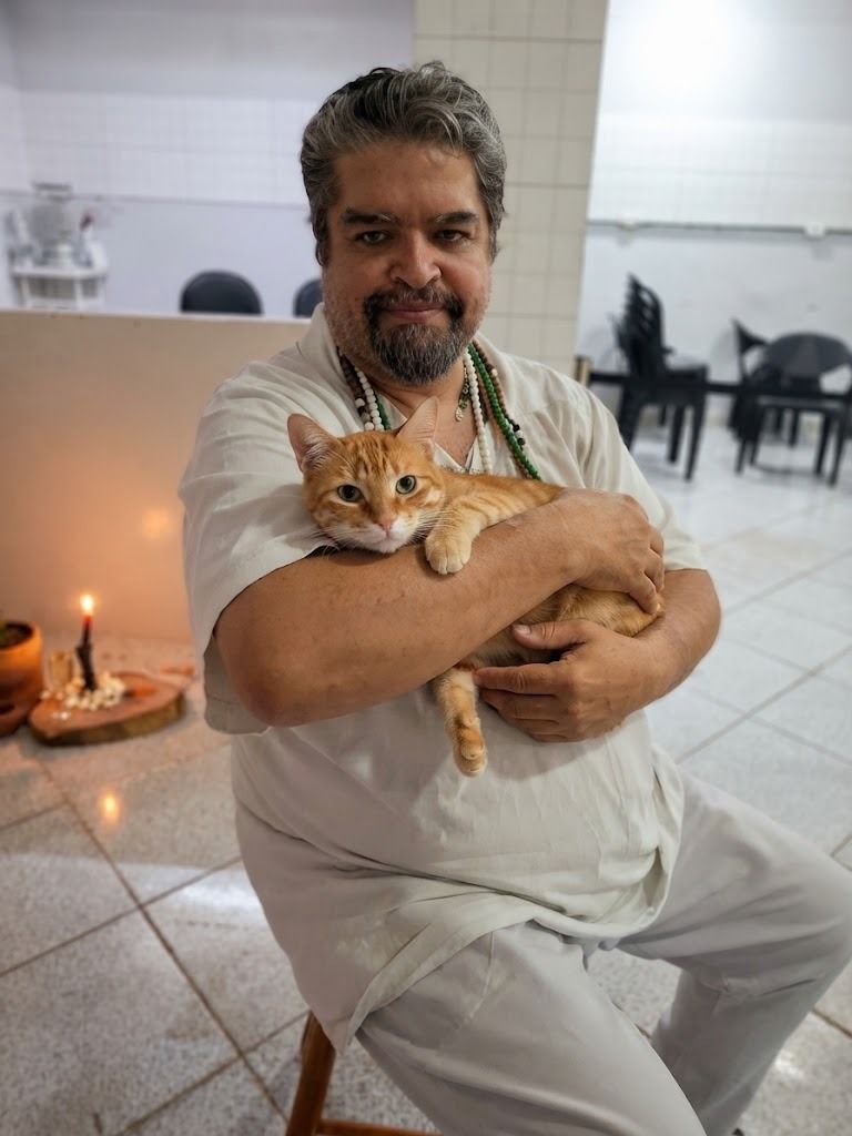 O dirigente do Templo, sentado e de roupa branca, segura no colo um gato laranja diante de uma vela acesa e um ponto de ervas.