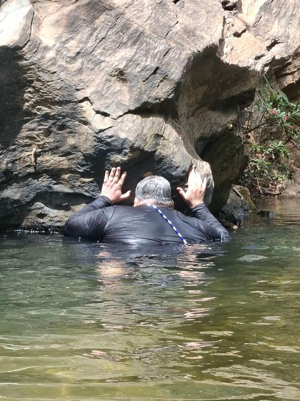 Com a água pela cintura, um médium de guia azul e branca apoia as mãos numa grande pedra à margem do Ribeirão dos Bois.