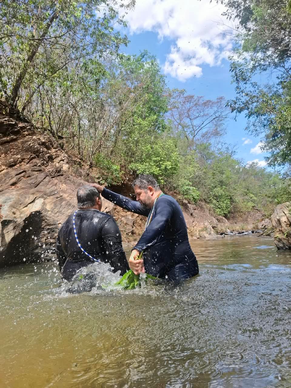 Dentro do Ribeirão dos Bois, um médium com um feixe de folhas na mão pousa a outra mão sobre a cabeça de outro médium durante o sacudimento.